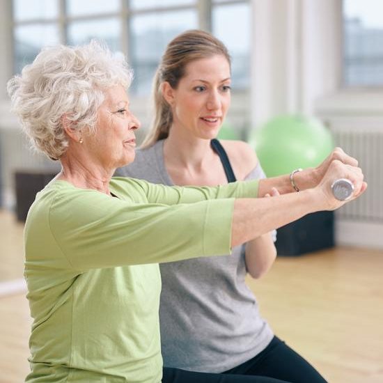 Senior woman being assisted by instructor in lifting dumbbells at gym. Senior woman training in the gym with a personal trainer at rehab.