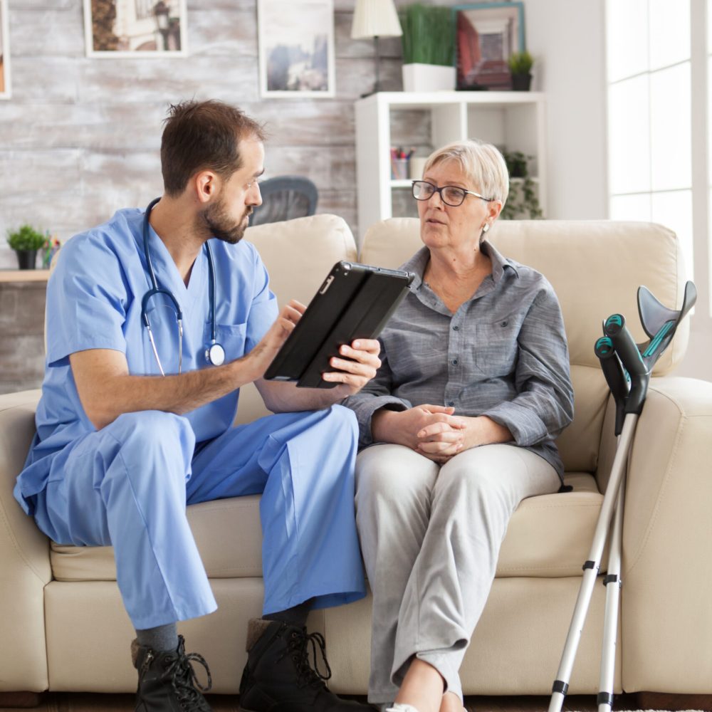 Old woman with crutches in nursing home sitting on couch listening male doctor with tablet computer.